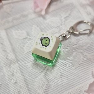 May include: A keycap keyring featuring a light green frog design on a cream-coloured square keycap. The keycap sits atop a transparent green base, attached to a silver-coloured keyring. The frog illustration is outlined in black.