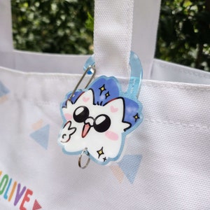 May include: A white tote bag with a blue and white cartoon character charm attached to the handle. The charm features a white cat with black eyes and a pink nose, wearing sunglasses and a blue hat. The charm is attached to the bag with a silver clip.