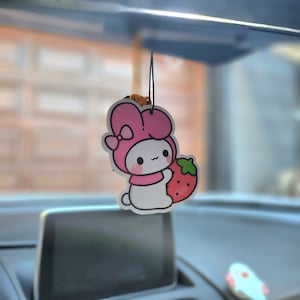 Puede incluir: Un personaje de dibujos animados rosa y blanco con un lazo sosteniendo una fresa roja. El personaje es un ambientador de coche.
