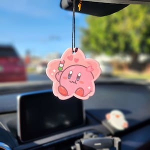 May include: Pink star-shaped car air freshener with a cartoon character holding a skewer of food. The character has pink skin, large eyes, and a small mouth. The air freshener is hanging from a rearview mirror.
