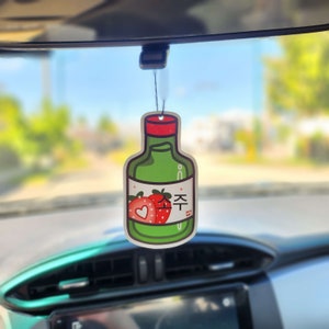 Puede incluir: Un ambientador verde con forma de botella de soju, con tapa roja y etiqueta con fresas y texto coreano. Cuelga del espejo retrovisor de un coche, con fondo borroso.