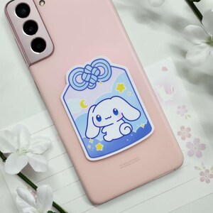 May include: A blue and white sticker featuring a cute cartoon bunny rabbit with a blue bow on a light blue background with stars and a crescent moon. The sticker is on a pink phone case.