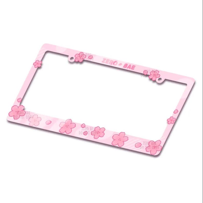 May include: A pink license plate frame adorned with pink cherry blossom flowers. The frame features the text "ZERO + BAE" and is designed to fit a standard license plate. The frame is made of a durable material.