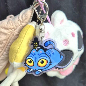 May include: A blue cat-shaped keychain with yellow eyes and a black hat. The keychain is attached to a silver clasp and a pink loop, with a white plush toy in the background.