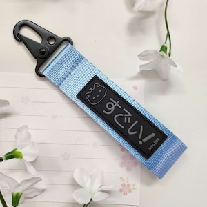 Può includere: Un portachiavi blu chiaro con un ciondolo a forma di gatto nero e il testo giapponese "すき!" (suki!), che significa "Mi piaci!" in inglese. Il portachiavi ha un gancio in metallo nero.