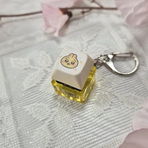 May include: A cream-colored keycap with a cartoon dumpling face, attached to a clear yellow resin base and a silver keychain. The keycap has a cute, smiling face with pink cheeks and small hearts. The background is a white lace fabric with pink flower accents.