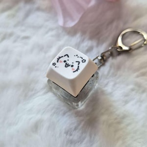 Peut inclure: Un porte-clés carré blanc avec un motif de visage de chat de dessin animé. Le porte-clés est attaché à un porte-clés argenté. Le fond est une surface douce et blanche avec quelques fleurs roses.