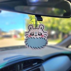 May include: A white and blue car air freshener shaped like a hamster with a fluffy blue body and pink cheeks. The air freshener is hanging from the rearview mirror of a car.