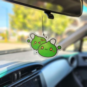 May include: A green car air freshener featuring two cartoon characters. The characters have large eyes and are smiling. The air freshener is hanging from the rear-view mirror.