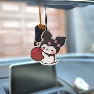 May include: A black and white car air freshener with a cartoon character holding a red cherry. The character has a pink bow and a black and white striped shirt. The air freshener is hanging from a string.