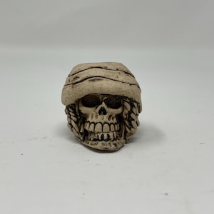 May include: A white ceramic skull with a brown fabric covering the top of the head. The skull has a detailed face with a toothy grin.
