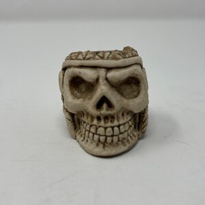 Può includere: Un piccolo teschio in ceramica bianca con un viso dettagliato e una corona di foglie. Il teschio ha un'espressione minacciosa con occhi infossati e un ghigno pieno di denti.