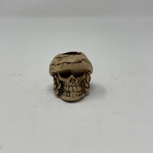 May include: A small, beige skull figurine with a detailed face and a textured, cracked top. The skull is wearing a hat.