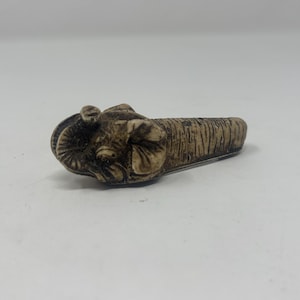 May include: A small, carved, ivory-colored elephant figurine. The elephant is lying on its side with its trunk curled up. The figurine is detailed with intricate carvings.