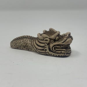 May include: A small, white ceramic dragon figurine with a detailed design. The dragon is curled up with its head raised and its mouth open.
