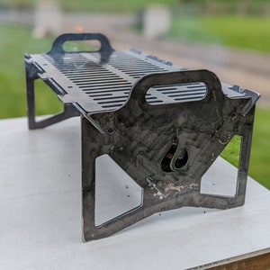May include: A small, portable metal grill with a slotted cooking surface and integrated handles. The grill has a flame-shaped cutout on the side and is supported by angled legs. It is designed for outdoor cooking and measures approximately 12 inches long.