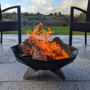 May include: A black metal fire pit with a geometric design, filled with burning logs and bright orange flames. The fire pit is on a stone patio, with two black chairs in the background. Green hills and a blue sky are visible.