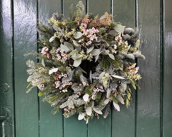 May include: A circular wreath composed of dark green pine needles, frosted evergreen branches, and silver-toned eucalyptus leaves. The arrangement is accented with clusters of small, light pink flowers and delicate, pale yellow floral sprigs.