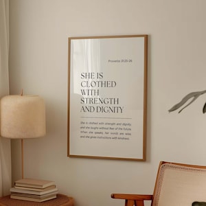 May include: A framed print with the text "SHE IS CLOTHED WITH STRENGTH AND DIGNITY" in a minimalist style. The print is in a light brown wooden frame and is hanging on a beige wall. A beige lamp and a small side table are in the foreground.