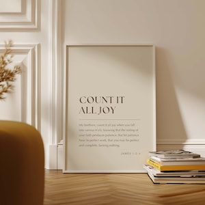 May include: A framed print with the text "COUNT IT ALL JOY" in a minimalist style. The print includes a Bible verse from James 1:2-4. The frame is white, and the background is a neutral beige color. A stack of books rests on the floor.