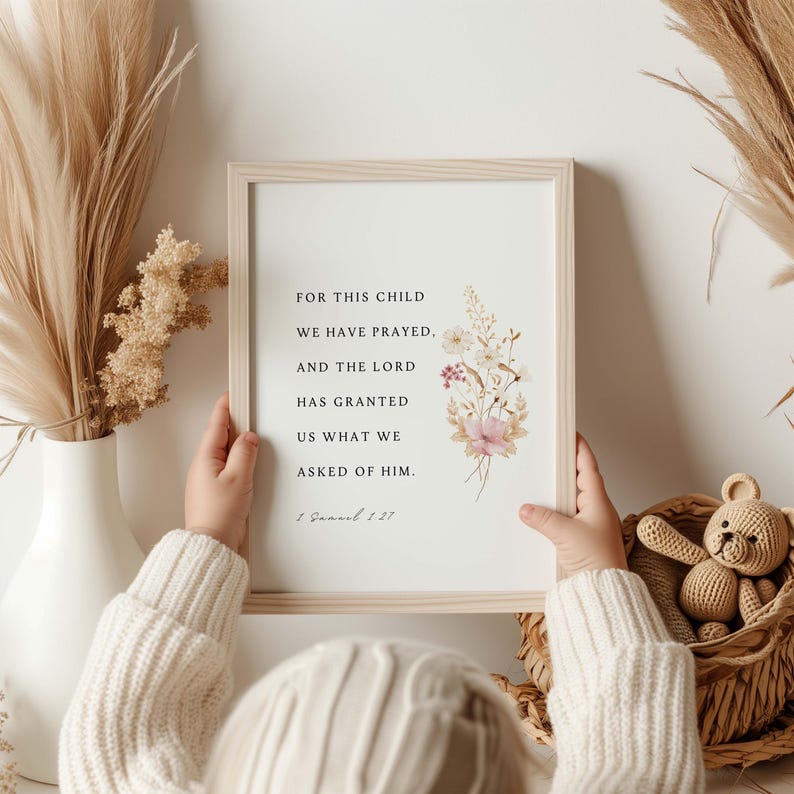 May include: A framed print with the text "FOR THIS CHILD WE HAVE PRAYED, AND THE LORD HAS GRANTED US WHAT WE ASKED OF HIM." Floral illustrations in pink and white adorn the right side. The frame is held by a person wearing a cream-colored sweater.