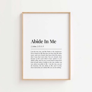 Op de afbeelding: Een ingelijste print met de tekst "Abide In Me" in zwart op een witte achtergrond. De print heeft een houten lijst en bevat een Bijbelvers uit Johannes 15:1-5. Een eenvoudig, elegant ontwerp.