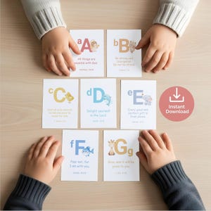 Könnte beinhalten: Alphabet-Lernkarten mit Illustrationen und Bibelversen. Jede Karte zeigt einen Buchstaben, ein Bild und ein Zitat. Die Karten liegen auf einer hellen Holzoberfläche, mit Kinderhänden sichtbar.