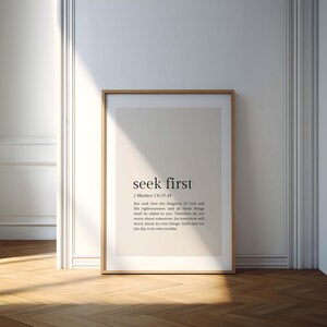 May include: A framed print with the words "seek first" in a minimalist design. The text is in black against a light beige background. The frame is a light brown color, and the print is leaning against a white wall and a wooden floor.