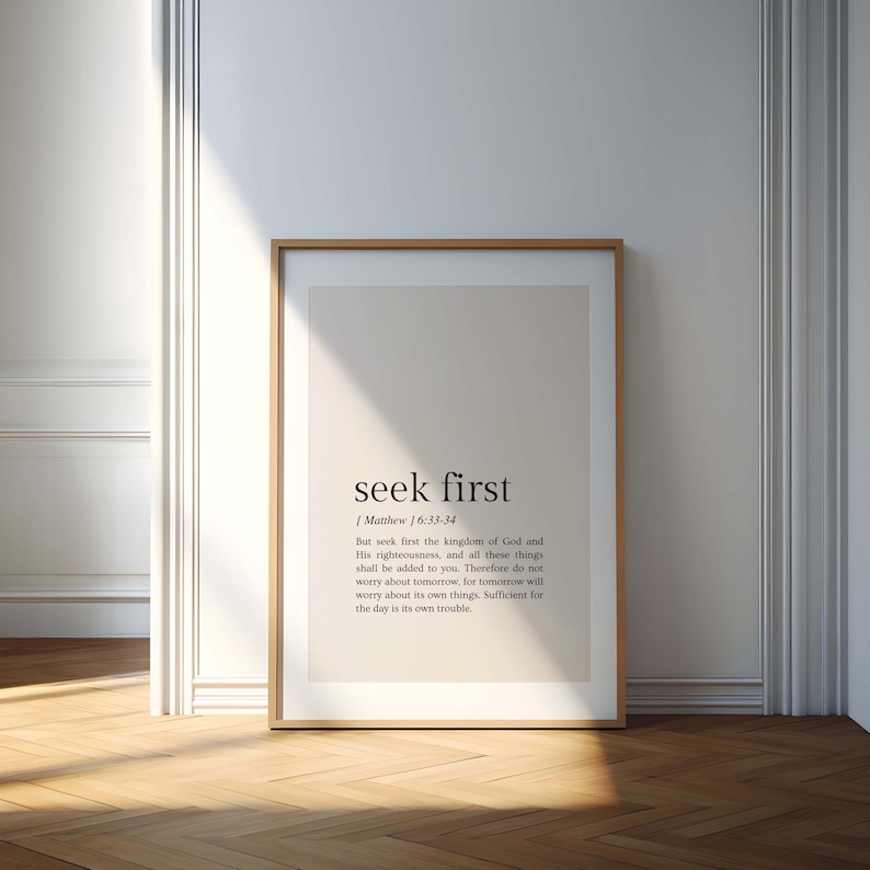 May include: A framed print with the words "seek first" in a minimalist design. The text is in black against a light beige background. The frame is a light brown color, and the print is leaning against a white wall and a wooden floor.
