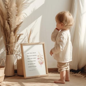 May include: A framed print with the text "FOR THIS CHILD WE HAVE PRAYED, AND THE LORD HAS GRANTED US WHAT WE ASKED OF HIM." features floral accents in pink and white. A young child stands nearby, wearing a cream-colored outfit.