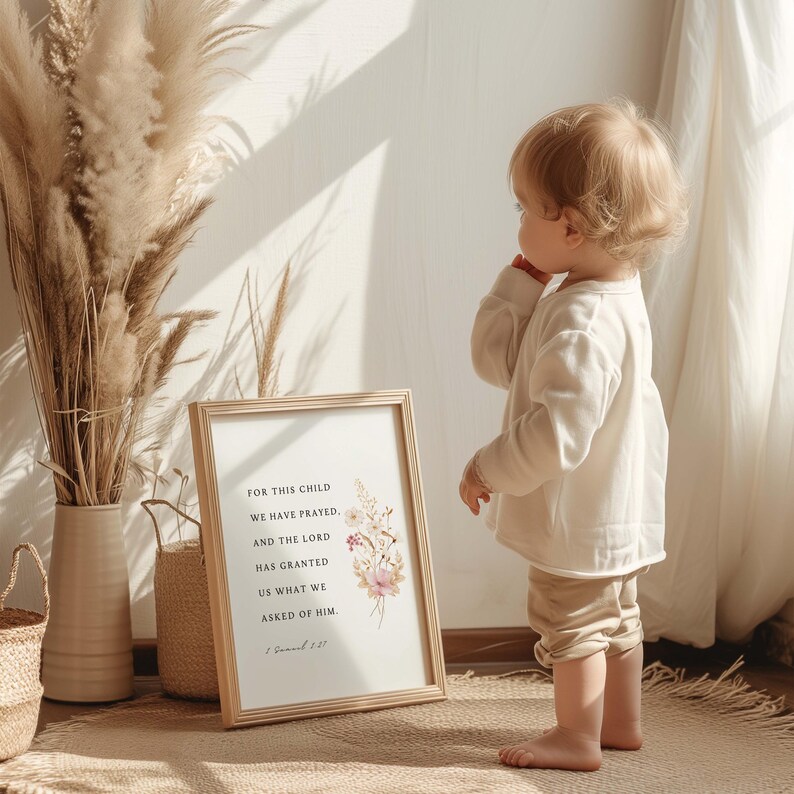 May include: A framed print with the text "FOR THIS CHILD WE HAVE PRAYED, AND THE LORD HAS GRANTED US WHAT WE ASKED OF HIM." features floral accents in pink and white. A young child stands nearby, wearing a cream-colored outfit.
