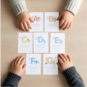 Può includere: Un set di sette schede dell'alfabeto con versetti biblici, con illustrazioni di un angelo, un orso, un pulcino, un delfino, un elefante, un pesce e una giraffa. Le schede sono disposte su una superficie di legno chiaro, con mani di bambini.