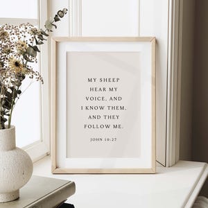 May include: A framed print with the text "MY SHEEP HEAR MY VOICE, AND I KNOW THEM, AND THEY FOLLOW ME. JOHN 10:27." The frame is light wood, and the background is a soft beige. A vase with dried flowers is to the left.