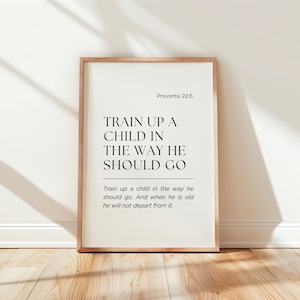 Op de afbeelding: Een ingelijste print met de tekst "TRAIN UP A CHILD IN THE WAY HE SHOULD GO" uit Spreuken 22:6. De print heeft een licht houten frame en wordt weergegeven op een houten vloer tegen een witte muur.