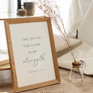 May include: A framed print with a white background and black text that reads "The joy of the Lord is my strength, Nehemiah 8:10". The frame is made of light brown wood and is sitting on a woven rug.