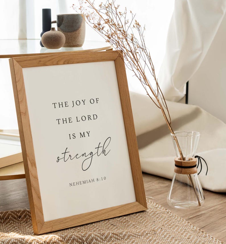May include: A framed print with a white background and black text that reads "The joy of the Lord is my strength, Nehemiah 8:10". The frame is made of light brown wood and is sitting on a woven rug.