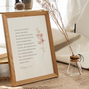 May include: A framed print with a light brown wood frame and a white background. The print features a quote from Philippians 4:6-7, "Do not be anxious about anything, but in every situation, by prayer and petition, with thanksgiving, present your requests to God. And the peace of God, which transcends all understanding, will guard your hearts and your minds in Christ Jesus." The quote is surrounded by a watercolor illustration of pink flowers.
