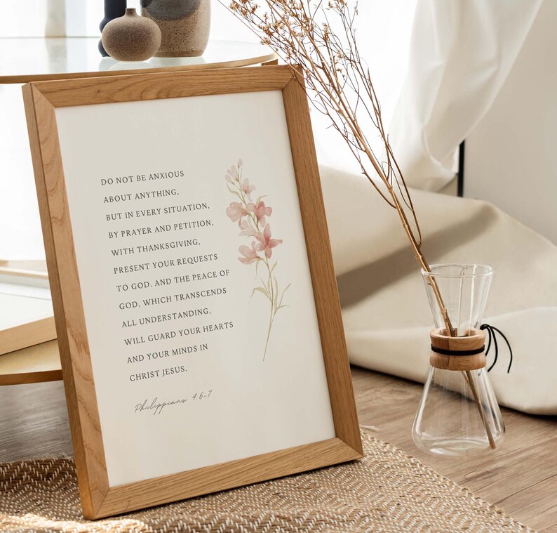 May include: A framed print with a light brown wood frame and a white background. The print features a quote from Philippians 4:6-7, "Do not be anxious about anything, but in every situation, by prayer and petition, with thanksgiving, present your requests to God. And the peace of God, which transcends all understanding, will guard your hearts and your minds in Christ Jesus." The quote is surrounded by a watercolor illustration of pink flowers.