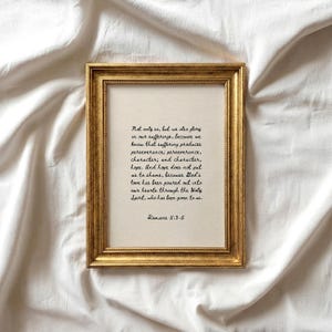 Puede incluir: Un marco dorado adornado enmarca una impresión con texto manuscrito de Romanos 5:3-5 sobre un fondo crema. El marco rectangular descansa sobre una superficie blanca texturizada.