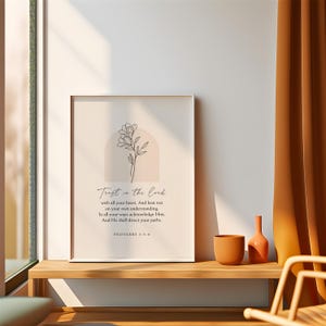 May include: A framed print with the text "Trust in the Lord" and a floral illustration. The artwork features a minimalist design with a beige background and a white frame. The print is displayed on a wooden shelf, alongside a terracotta vase and a small orange bottle.