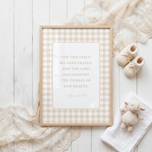 Può includere: Una stampa incorniciata con un bordo a quadretti beige e il testo "FOR THIS CHILD WE HAVE PRAYED, AND THE LORD HAS GRANTED THE DESIRES OF OUR HEARTS. 1 Samuel 1:27". L'immagine include scarpine per bambini, un peluche di agnello e un panno di mussola bianco.