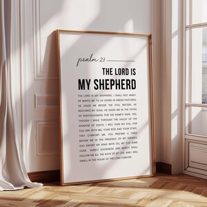 May include: A framed print with the text of Psalm 23, featuring the words "The Lord is My Shepherd" in bold. The print is in a gold frame and leans against a white wall and wooden floor, creating a warm, inviting aesthetic.