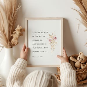 May include: A rectangular wooden frame containing a white print with the black text Train up a child in the way he should go, and when he is old he will not depart from it. Proverbs 22:6, decorated with a small watercolor floral illustration.