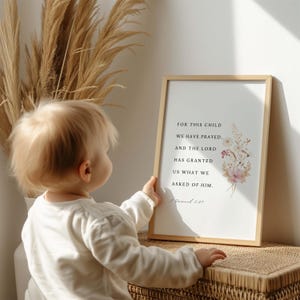 May include: A framed print with the text "FOR THIS CHILD WE HAVE PRAYED, AND THE LORD HAS GRANTED US WHAT WE ASKED OF HIM." The print features a floral design in soft pinks and whites. A baby is looking at the framed print.