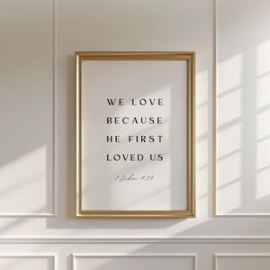 Może przedstawiać: Oprawiony wydruk z tekstem "WE LOVE BECAUSE HE FIRST LOVED US" i odniesieniem do Biblii 1 Jana 4:19. Rama jest złota, a tekst jest prostą, elegancką czcionką.