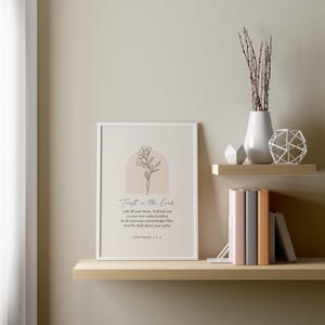 May include: A framed print with the text "Trust in the Lord" and a floral illustration. The print is displayed on a wooden shelf, alongside books, a vase with branches, and decorative objects. The color palette is neutral with beige, white, and brown tones.