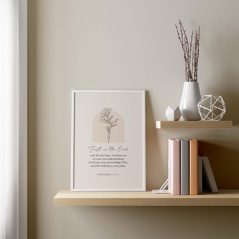 May include: A framed print with the text "Trust in the Lord" and a floral illustration. The print is displayed on a wooden shelf, alongside books, a vase with branches, and decorative objects. The color palette is neutral with beige, white, and brown tones.