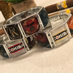 May include: Three silver charm bracelets with different cigar brand logos. The bracelets feature silver metal links and square glass charms with colorful images of cigar brands, including "Punch Clasico", "Cohiba Connecticut", "Romeo y Julieta", "Cohiba Royale", and "Sharer Million & Solomon".