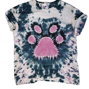 May include: A tie-dye t-shirt featuring a pink paw print design. The shirt is a light blue colour with dark blue and white tie-dye patterns.