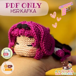 Op de afbeelding: Een roze en zwarte gehaakte amigurumi pop met gesloten ogen. De pop ligt op een hand. De afbeelding bevat de tekst "PDF ONLY HSR KAFKA".